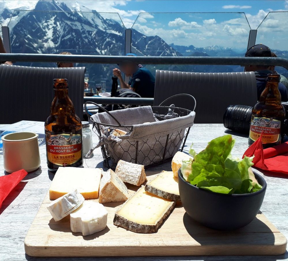 Caption: A photo of a cheese plate and bottles of beer at Le Panoramic Mont Blanc in Chamonix, France, an outdoor restaurant at the summit of Brévent, with stunning views of Mont Blanc in the background. (Local Guide Katalin Lippa)