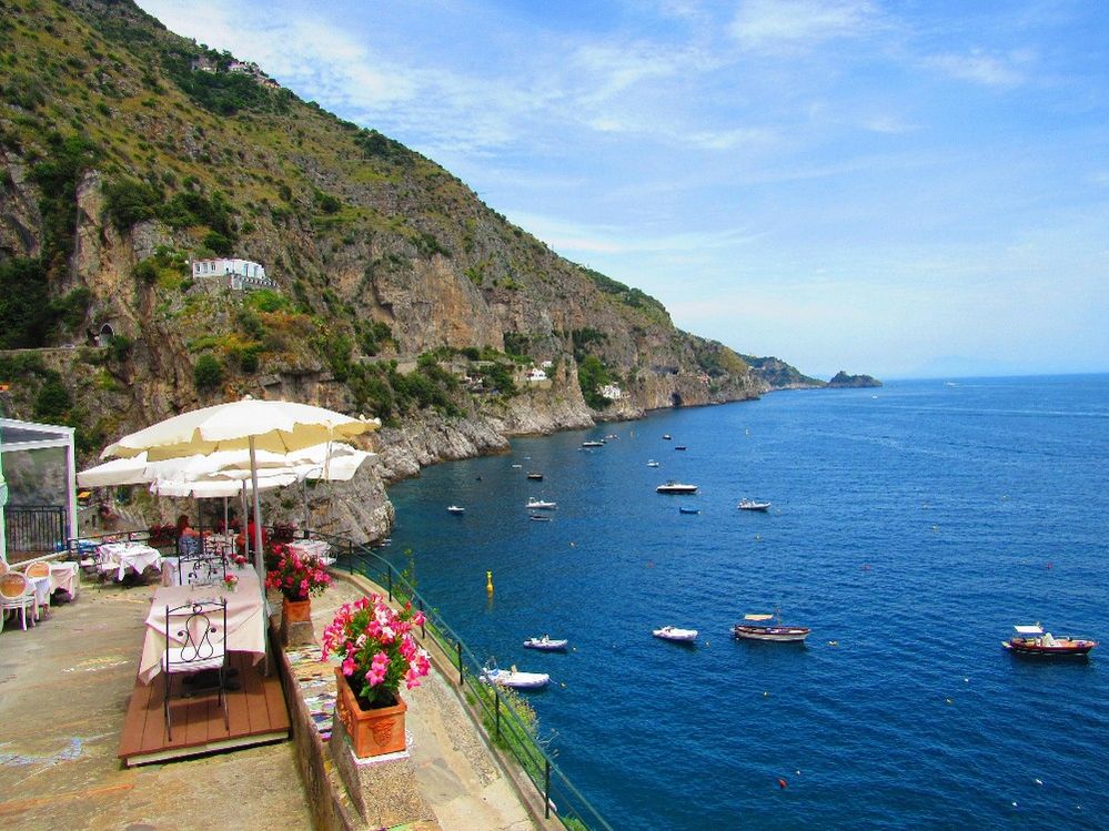 Caption: A photo of restaurant tables on a patio under umbrellas at Il Pirata on the Amalfi Coast overlooking the sea and some boats with cliffs in the background. (Local Guide Tom Trombino)