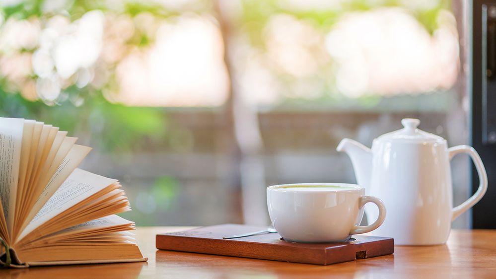 Caption: A photo of an open book, a white teapot, and a white coffee cup on a wooden tray next to a spoon on a table. (Getty Images)