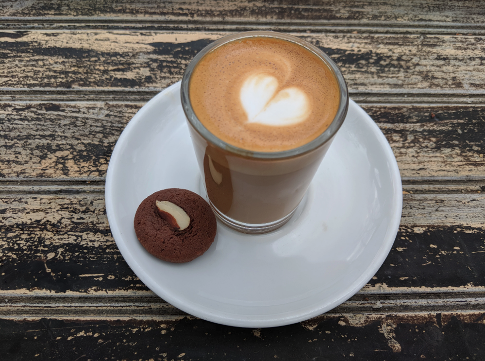 Caption: A photo of a coffee drink in a glass cup with a heart shaped design made in foam on a saucer next to a cookie. (Local Guide Daniel Ngo)