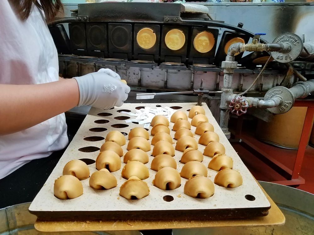 Caption: A person filling a fortune cookie with a paper fortune over a tray filled with more fortune cookies at the Golden Gate Fortune Cookie Factory in San Francisco, California. (Local Guide Frank Jang)