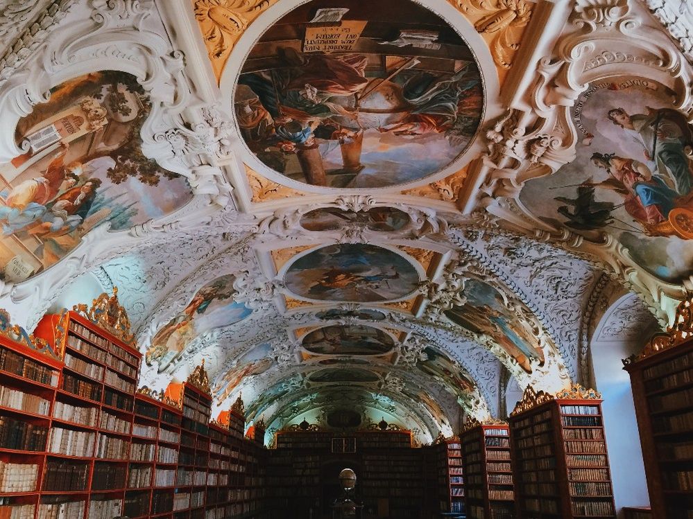 Caption: A photo of the Baroque Library at The Clementinum, a historic site in Prague, Czech Republic. The ornate ceiling is decorated with frescoes that depict Jesuit saints. Bookshelves line the walls and a globe can be seen in the distance. (Local Guide TheHeuteCher)
