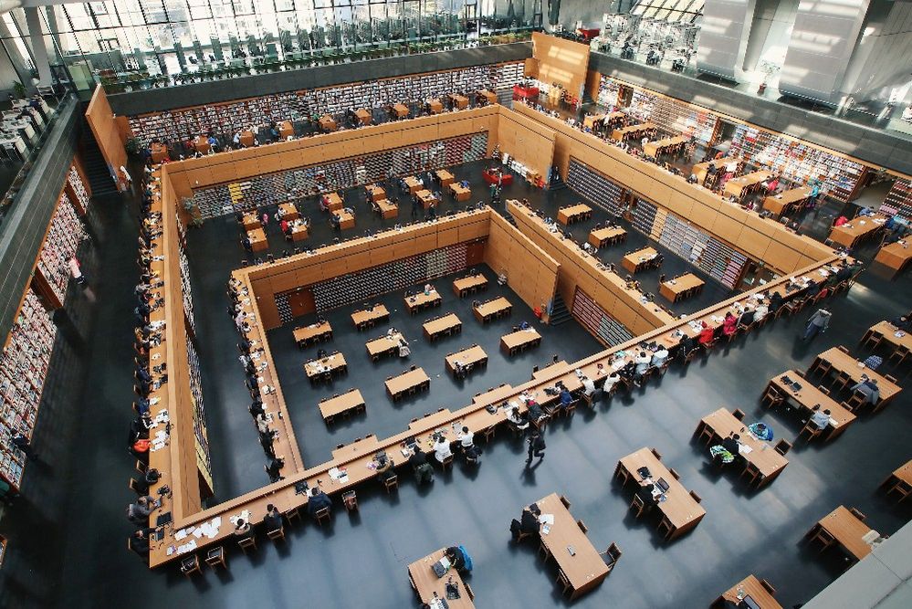 Caption: A photo of the interior of The National Library of China in Beijing, China that shows multiple levels filled with wooden tables with people reading at them. A large square shape is cut out of the center of each floor so that you can see the level below it. The walls of each level are lined with bookshelves. (Local Guide Kaiji Sun)