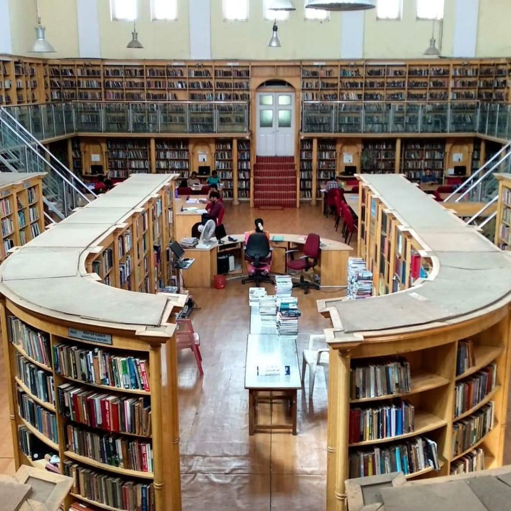 Caption: A photo of the interior of the State Central Library in Karnataka, India that shows two large, curved bookshelves in the center of the space. In between them, there are tables and a front desk for those who work there to sit. The walls of the space are filled with additional wall to wall bookshelves. (Local Guide Mayur Panchamia)