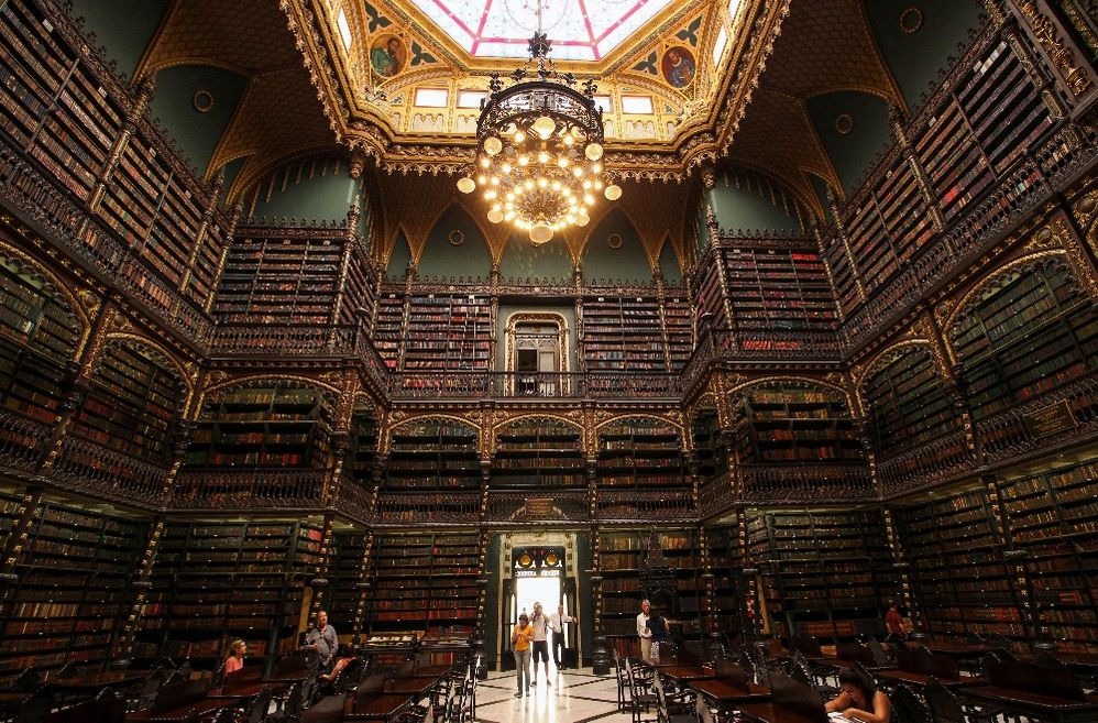 Caption: A photo of the interior of Royal Portuguese Cabinet of Reading, a library in Rio de Janeiro, Brazil. The ornate library has dark wall to ceiling bookshelves, a large chandelier in the center of the room, and a skylight. (Renan William Candido)