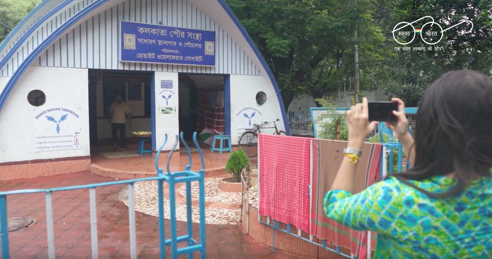 Caption: A photo of the back view of a woman in India taking a photo of the outside of a public toilet using her phone.
