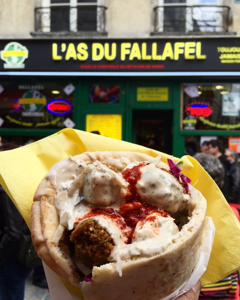 Caption: A photo of falafel in a pita with white sauce on top wrapped in a napkin and held up in front of the yellow sign and green storefront of L’As du Fallafel in Paris. (Local Guide Kristin Rimbach)