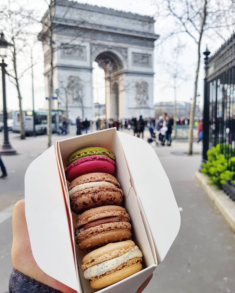 Caption: A photo of a small box of four different flavors of colorful macarons from Pierre Hermé in front of the Arc de Triomphe in Paris. (Local Guide Mehdi Idalene)