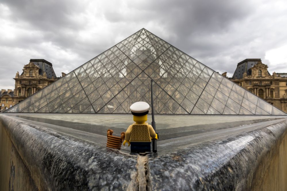 Caption: A photo of the back view of a LEGO minifigure wearing a hat and holding a fishing rod in front of the Louvre Pyramid at the Louvre Museum in Paris, France on a cloudy day. (Local Guide Julien Ballester)