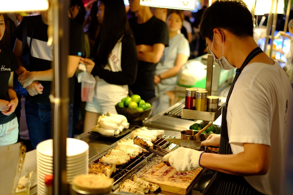 Caption: A photo of a man in an apron and a mask holding a pair of cooking tongs in front of a stall where food is cooking as a crowd lines up at Shilin Night Market in Taipei City, Taiwan. (Local Guide Natchapat Podee)