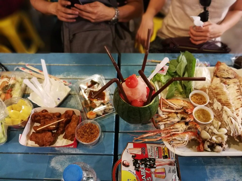 Caption: A photo of several containers of food and a tray with cooked fish and shrimp next to a drink with five straws served in a small carved-out watermelon. The food and drink are placed on a blue picnic table at Ratchada Rot Fai Night Market in Bangkok. (Anna Camille Ramirez)