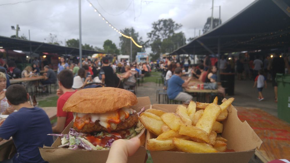Caption: A photo of a cheeseburger and French fries in a cardboard container held up in front of the many picnic tables and string lights at Mt Gravatt Marketta in Mount Gravatt, Australia. (Local Guide Sos Mattsson)