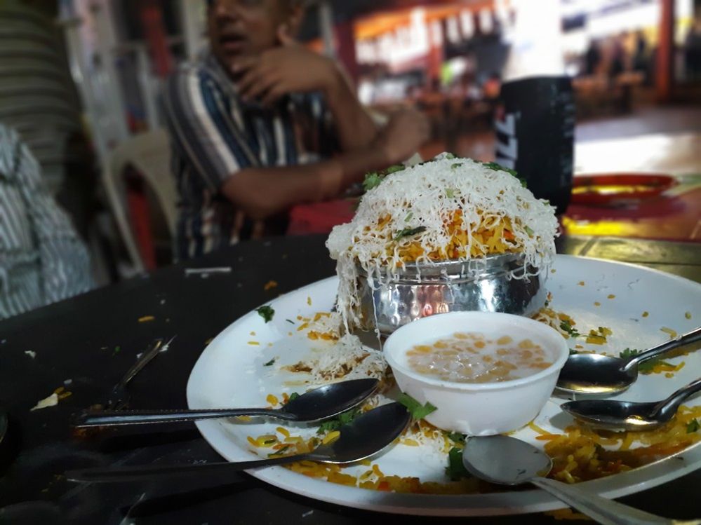 Caption: A photo of a local dish of rice with cheese and other toppings on a dish surrounded by spoons on a table at Ratri Bazaar. (Local Guide Jignesh Mahale)