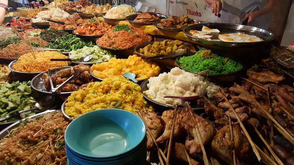 Caption: A photo of a street food stall at Night Market in Luang Prabang, Laos with platers of rice, noodles, fruit, vegetables, and more in addition to meat on skewers. (Local Guide Mam Sutheera)