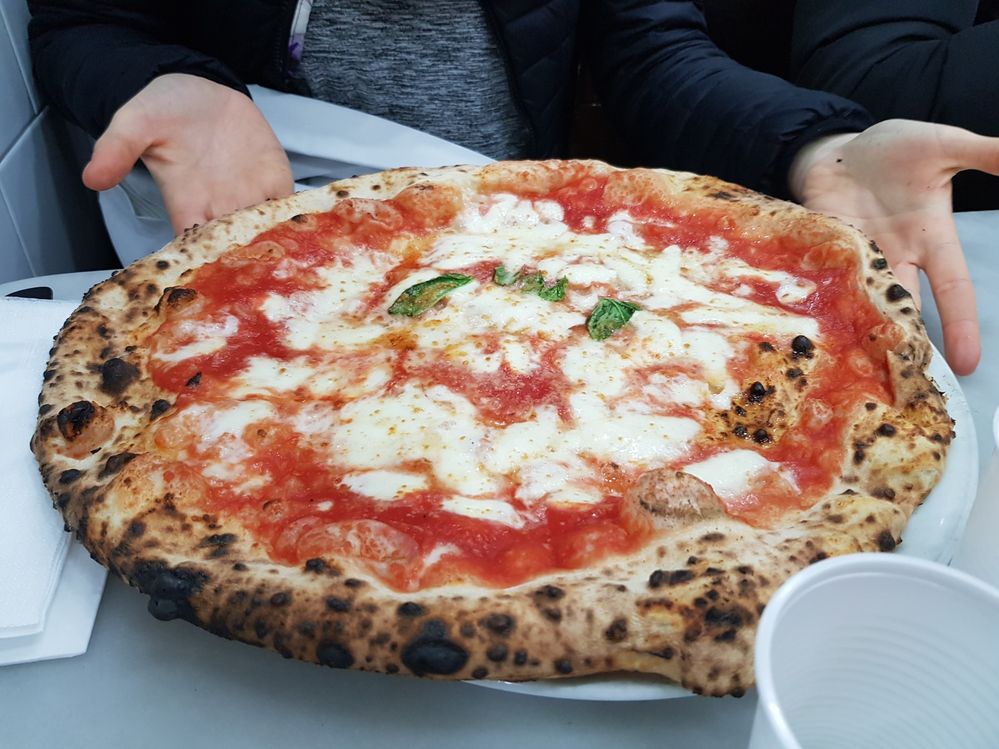 Caption: A photo of hands holding up a plate with a classic Margherita pizza pie on it at L’Antica Pizzeria da Michele in Napoli, Italy. The pie has mozzarella cheese, tomato sauce, and a few pieces of basil on it. (Local Guide Gianni Lupo)