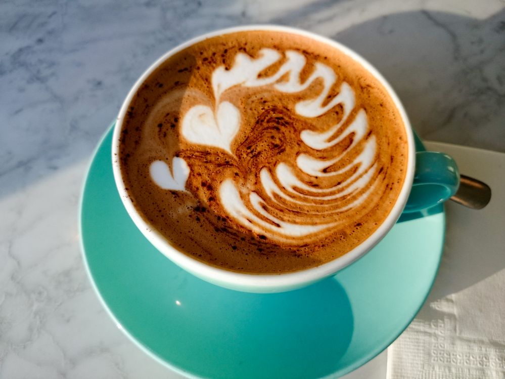 Caption: A photo of a latte topped with a design of hearts made with steamed milk in a turquoise cup with matching saucer on a marble table at 49th Parallel Café & Lucky's Doughnuts in Vancouver, Canada. (Local Guide E_____ C_____)