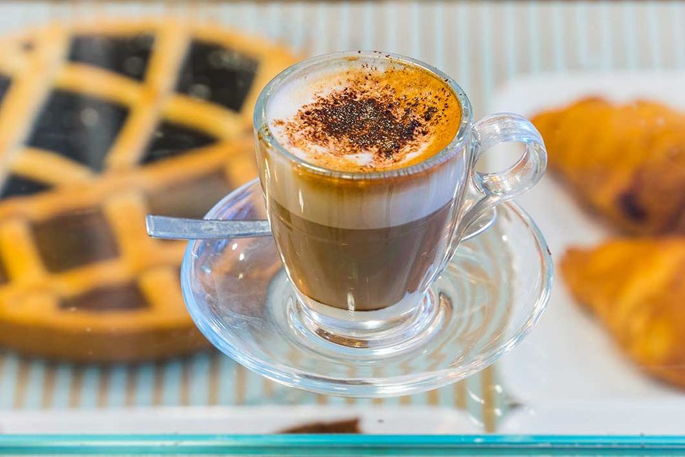 Caption: A photo of a coffee drink in a glass cup sitting on top of a pastry case containing a pie and two croissants. (Local Guide Massimo Pastori)
