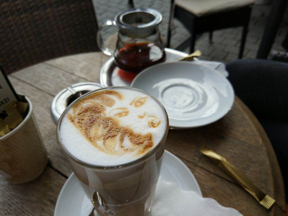 Caption: A photo of a coffee drink with a woman's face made in latte art on a wooden table next to a cup of sugar packets, a saucer, and a pot of coffee. (Local Guide Jennifer Pfeil)