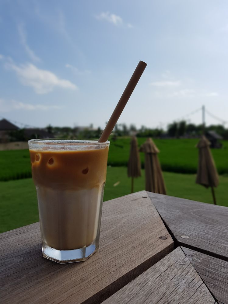 Caption: A photo of an iced coffee with a straw in a glass cup sitting on a wooden surface outdoors with grass, umbrellas, and a cloudy sky visible in the background. (Local Guide Danis Darusman)