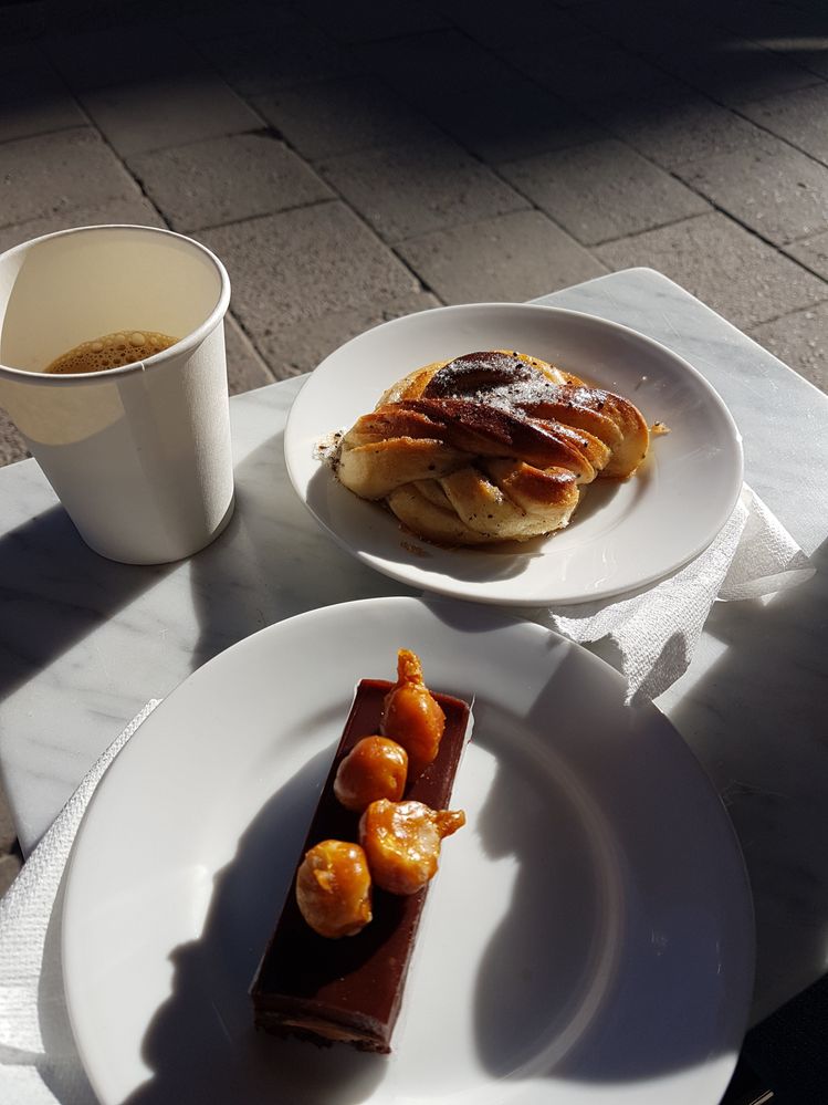 Caption: A photo of a Swedish cinnamon roll and small, rectangular piece of cake on plates on a white marble table outside at Lillebrors bageri in Stockholm, Sweden. (Local Guide Anna Joo)