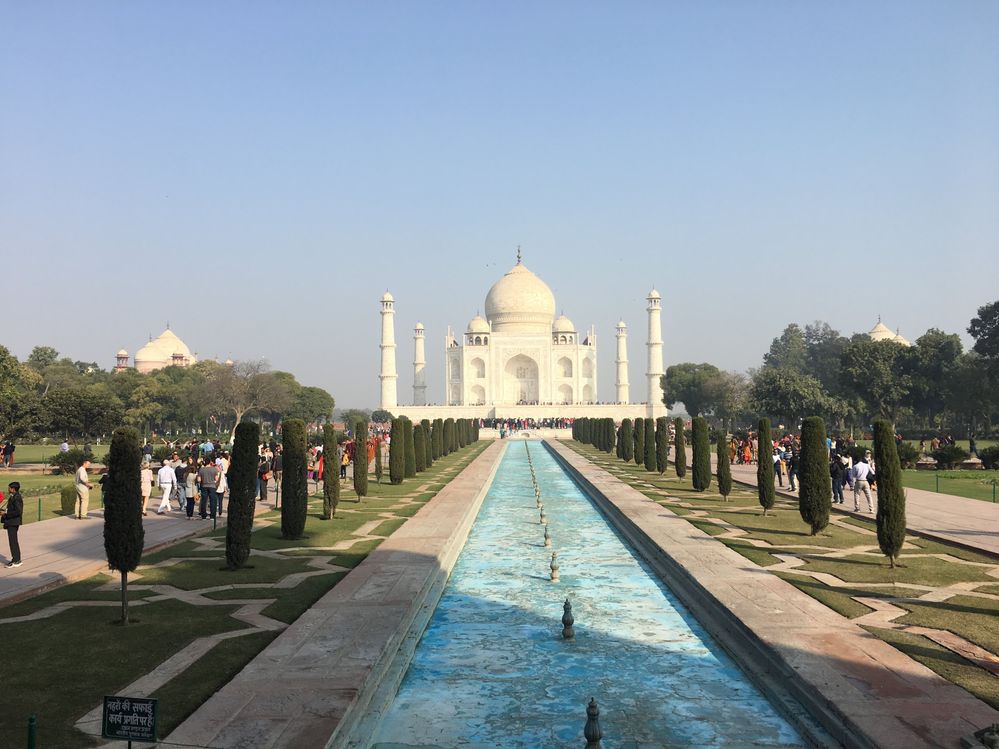 Caption: A photo of the Taj Mahal in Agra, India, showing the white marble mausoleum against a blue sky with its well-manicured garden and blue, dry fountain in front, and tourists filling the pathways on either side. (Local Guide Thanakorn Chokpitiboon)