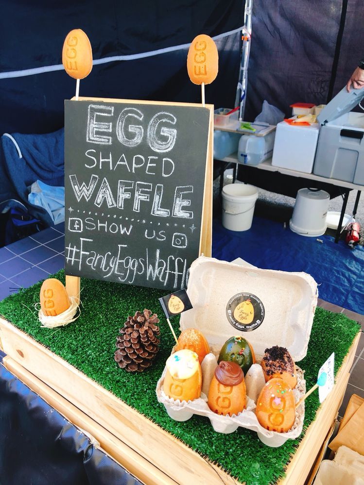 Caption: A photo of a vendor stand with a sign that says “Egg shaped waffle. Show us #FancyEggsWaffle” written in chalk next to a pinecone and a cardboard egg carton filled with six different egg-shaped waffle treats. (Local Guide @Sophievava)