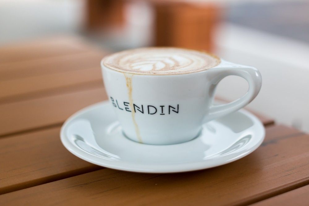 Caption: A photo of a coffee with a leaf design created in foam in a white ceramic cup and matching saucer on a wooden table at BlendIn Coffee Club in Sugar Land, Texas, USA. A streak of spilled coffee can be seen running down the cup. (Local Guide Daniel Ngo)