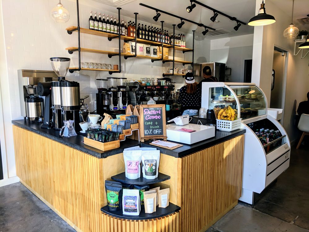 Caption: A photo of the counter at Luce Ave Coffee Roasters in Houston, Texas, filled with coffee equipment, tea, ground coffee beans for sale, and more. Behind the counter is even more equipment, shelves lined with syrups, coffee, and cups, and two people working with their backs turned. (Local Guide Daniel Ngo)