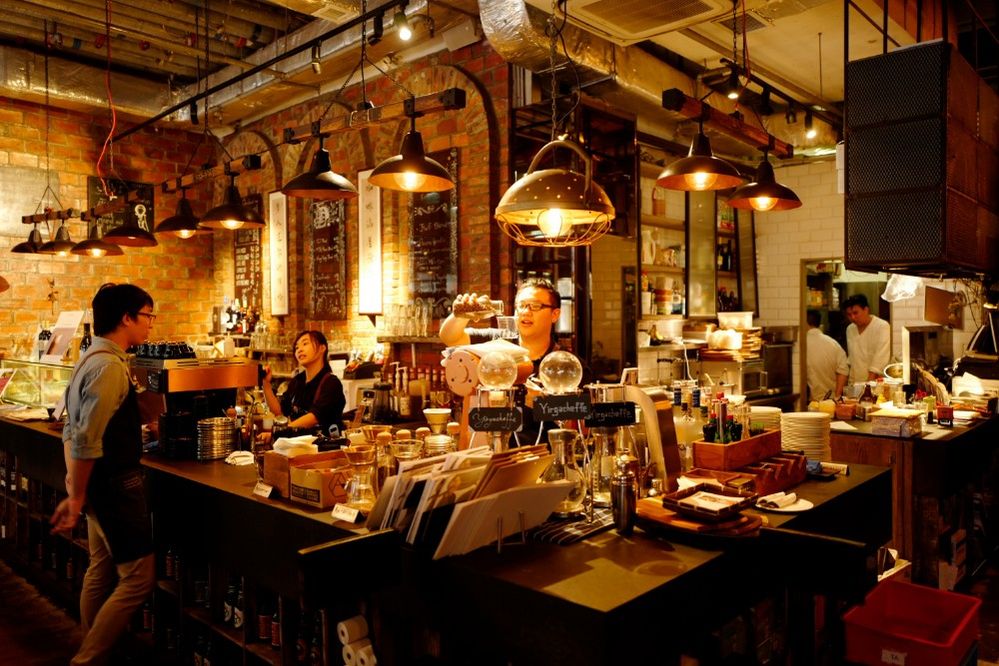 Caption: A photo of the interior of The Coffee Academics, a coffee shop in Hong Kong, showing baristas making drinks under the warm glow of the shop’s lights as well as the main counter that’s cluttered with coffee-making supplies like bean grinders, espresso machines, and coffee siphons. (Local Guide Jerry Tieng)