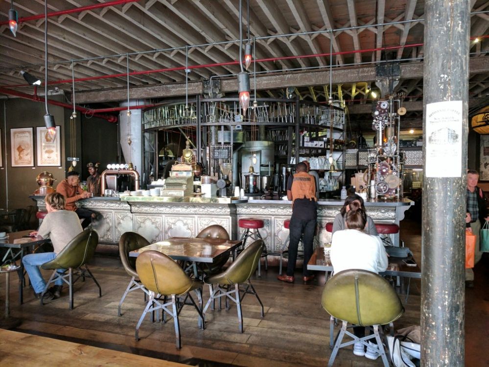 Caption: A photo of Truth Cafe, a steampunk-inspired cafe, in Cape Town, South Africa showing customers sitting at three metal tables with green chairs as well as the shop’s counter that’s decorated with metal pipes and old machinery. (Local Guide Chris Robinson)