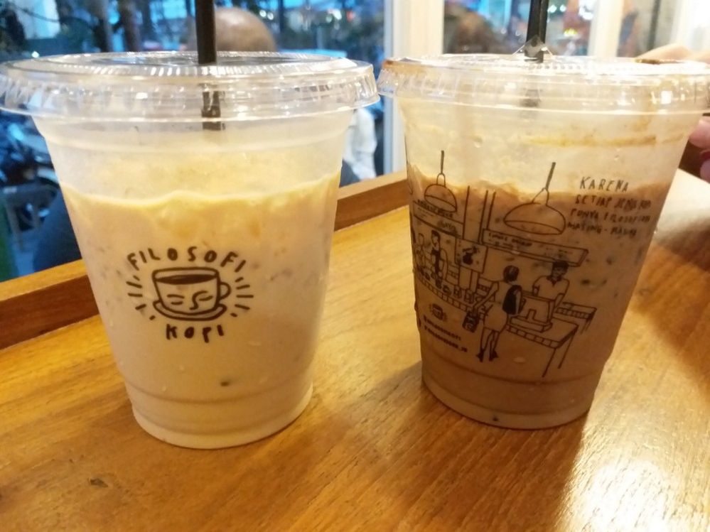 Caption: A photo of two iced drinks in plastic cups from Filosofi Kopi Melawai in Jakarta with illustrations on them depicting the coffee shop’s interior and its name. (Local Guide Aurelia Dessy)