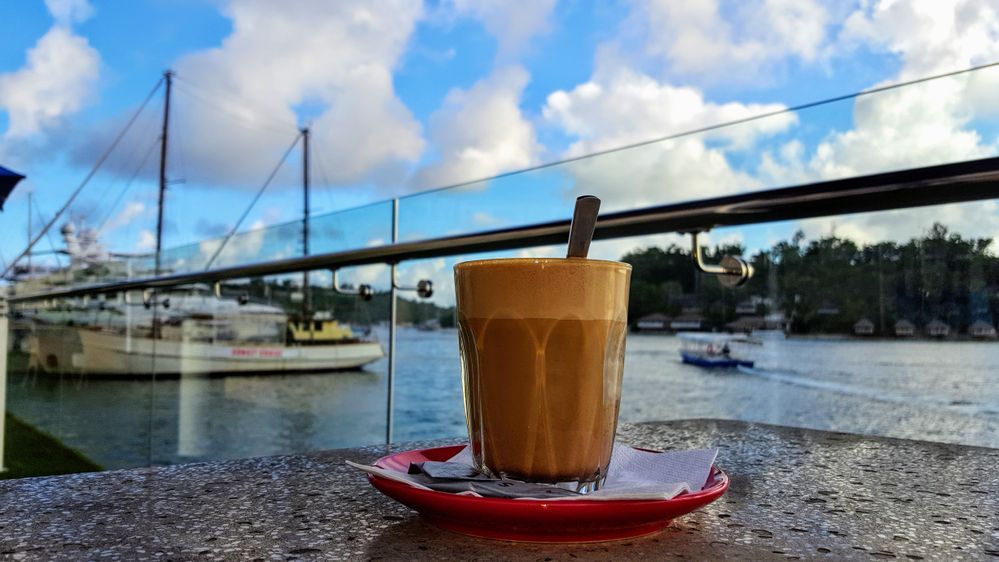 caption: A photo of a coffee drink in a glass cup on top of a white napkin on a small, red plate taken outside on a cloudy day at Grand Hotel and Casino in Port Vila, Vanuatu. Boats can be seen in the distance. (Local Guide Nick Hobgood)