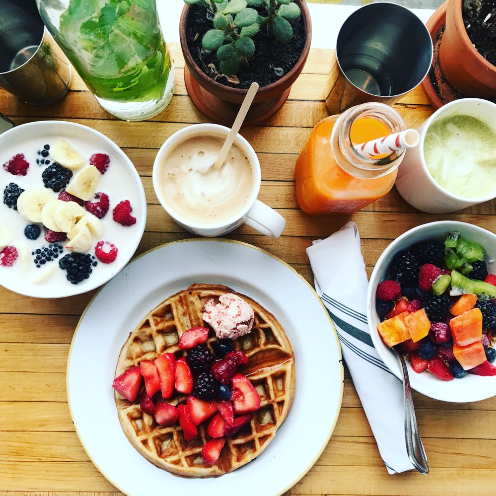 Caption: A photo of a Belgian waffle topped with berries, a bowl of fruit, a bowl of fruit with milk, a coffee, a matcha latte, a orange-colored juice, and a potted succulent on a wooden table at the Butcher’s Daughter in New York City. (Local Guide Mia Maria)