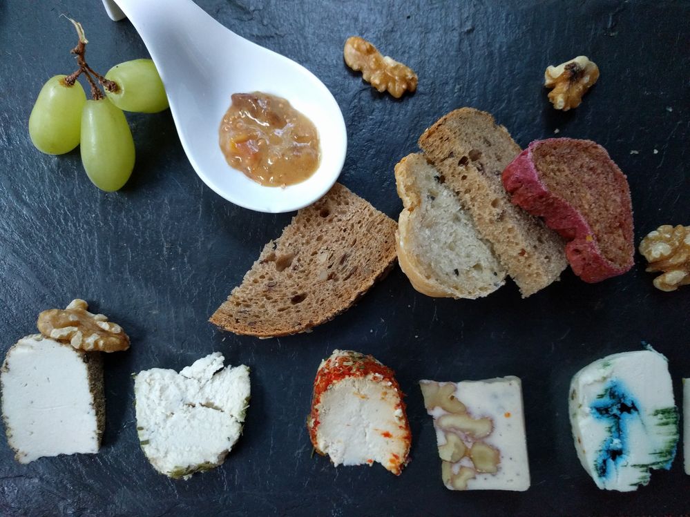 Caption: A photo of a slate cheese board topped with five types of vegan “cheeses” along with pieces of bread, nuts, grapes, and a type of jam in a large white spoon. (Local Guide Tobias Leenaert)