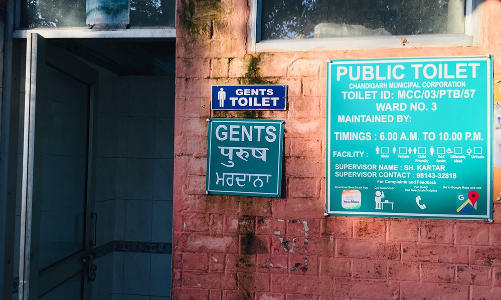 Caption: A photo of the outside of a public toilet in India. There’s two “Gents” signs and a “Public Toilet” sign with information like open hours and supervisor name. (Local Guide Emm Dee Singh)