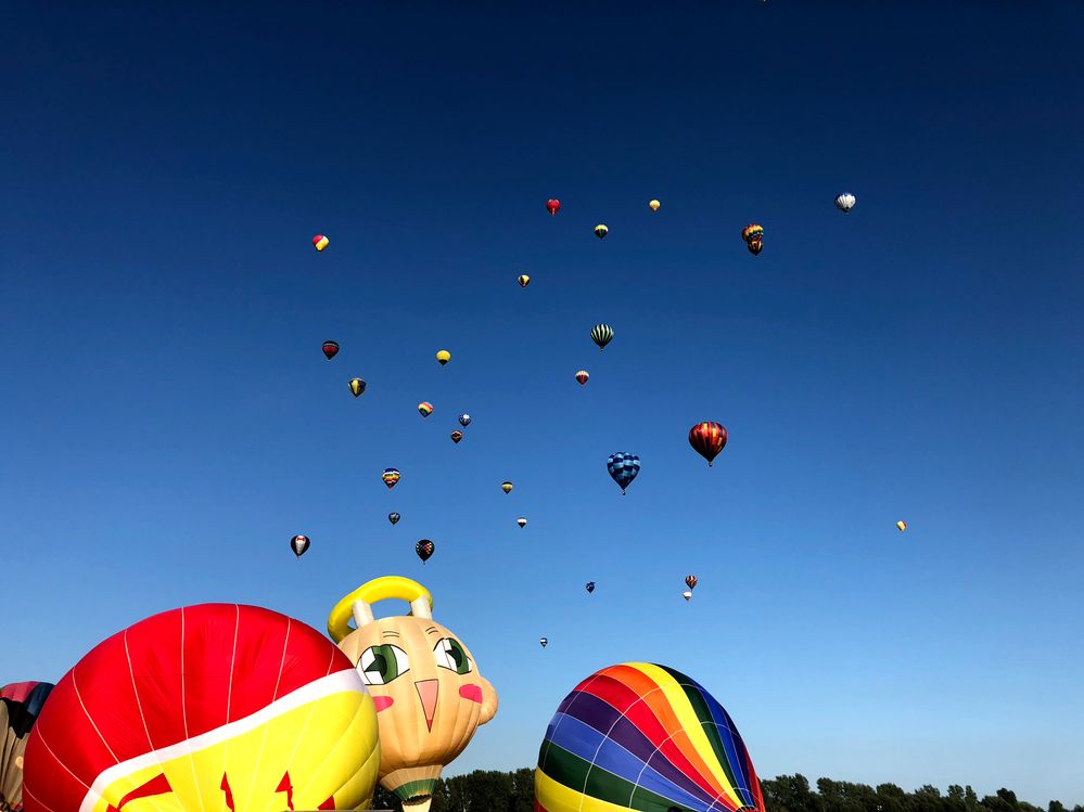Caption: A photo of a blue sky dotted with colorful hot air balloons in the distance and a few in the foreground, including one that looks like a cartoon face, at International de montgolfières de Saint-Jean-Sur-Richelieu in Quebec, Canada. (Local Guide Peter Galanopoulos)