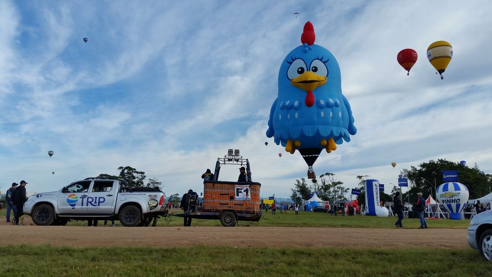 Caption: A photo of the blue sky with a blue hot air balloon that looks like a cartoon chicken flying and a few other hot air balloons in the distance, as well as a foreground of green grass, spectators, a pick-up truck hauling a hot air balloon basket on a strip of dirt road at Ballooning Park in Torres, Brazil. (Local Guide Ricardo Jardim)