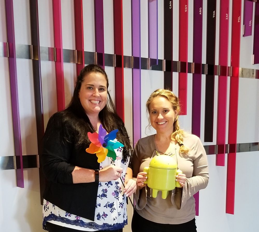 Caption: A photo of Connect Live 2018 event organizers Traci (left) holding a pinwheel and Brittany (right) holding a Local Guides Android figurine in front of a wall with multicolored stripes.