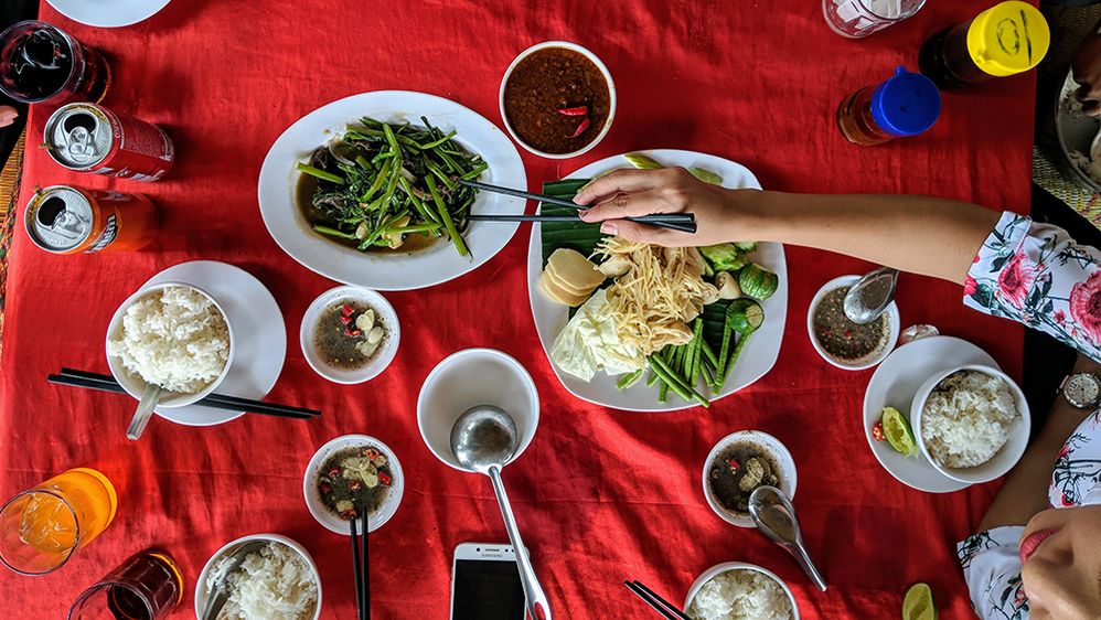 Caption: A photo of several plates of food on a red tablecloth taken from above at Cheng Meas Restaurant in Phnom Penh, Cambodia. A person’s hand can be seen reaching over to grab vegetables with chopsticks. (Local Guide Chamnan Muon)