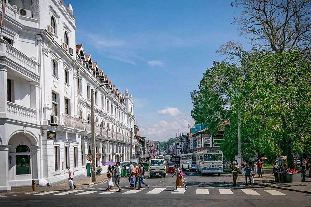 Caption: A photo of a street in Kandy, Sri Lanka. Pedestrians are crossing the street toward a large white building with many windows and balconies. (Local Guide Anuradha Piyadasa)