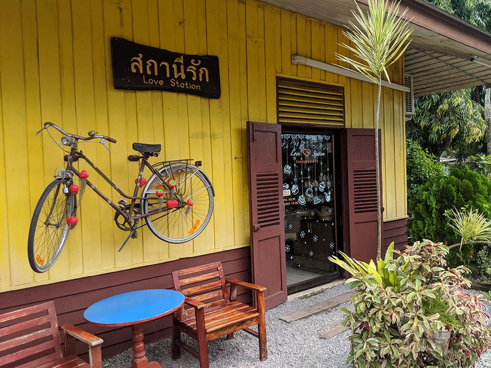 Caption: A photo of the outside of Love Station, a coffee shop in Trang, Thailand. A bicycle hangs underneath the coffee shop’s sign and above a small table with two wooden chairs beside it. (Local Guide Joseph Dewey)
