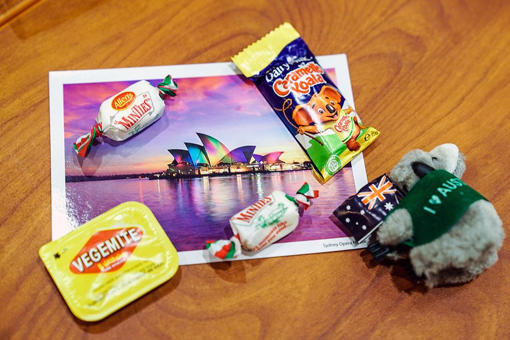 Caption: A photo of several items on a wooden table including a Cadbury caramel chocolate koala, two chewy peppermint Allen’s Minties, a mini tub of Vegemite, a small stuffed koala, and a photo of the Sydney Opera House.