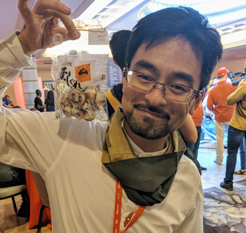Caption: A photo of Local Guide Hiroyuki holding up a bag of kuri shigare, a Japanese sweet treat made from chestnuts, at Connect Live 2018.