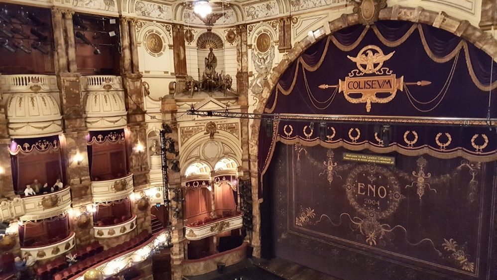 Caption: A photo of the ornate cream colored, gilded interior of the English National Opera in London with its plush red seats and large stage with a black and gold painted curtain. (Local Guide Sue Pyman)