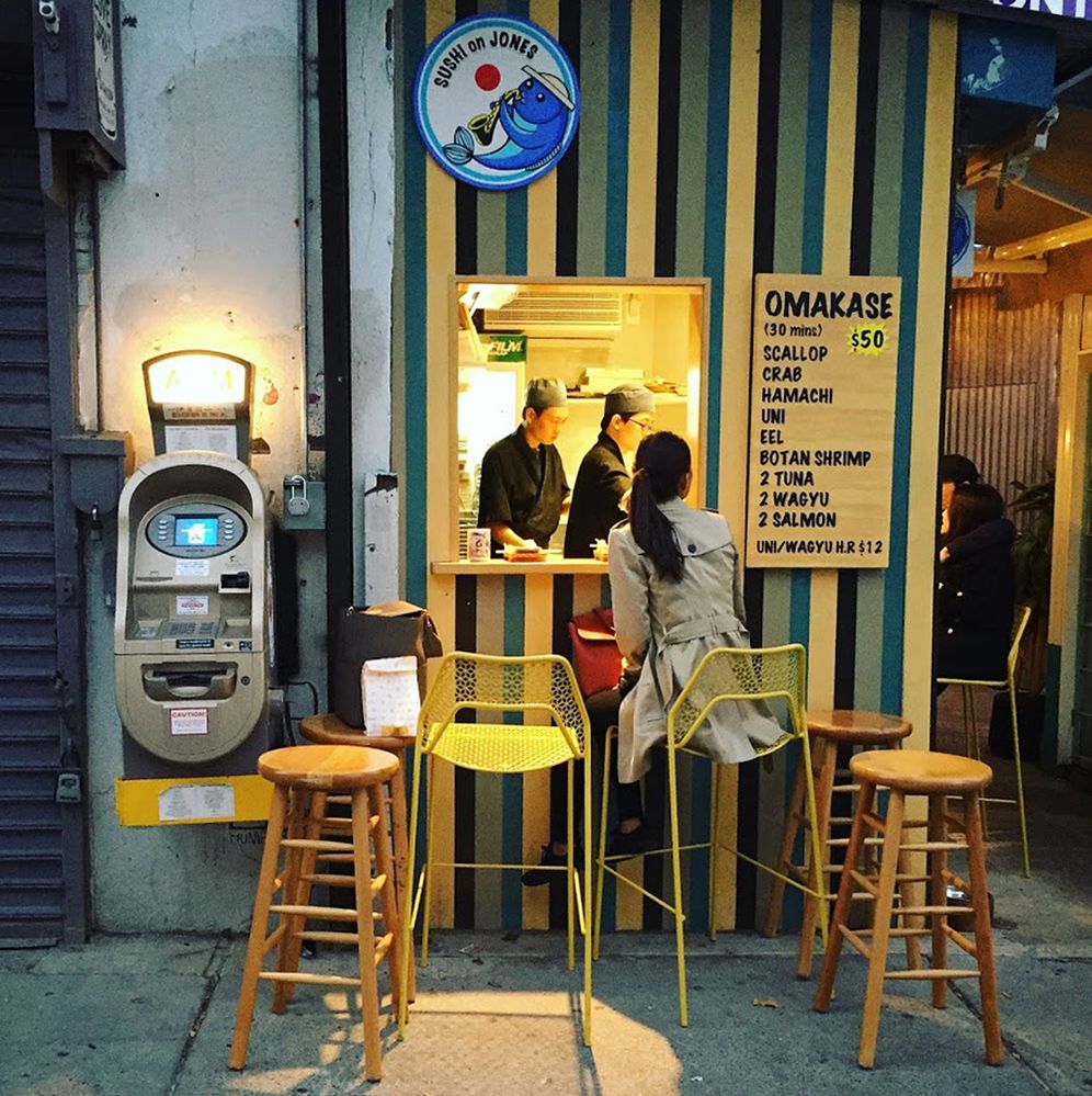 Caption: A photo of the exterior Sushi on Jones in New York, New York. A woman is sitting at the counter outside looking at the menu. She’s surrounded by chairs. (Local Guide Rohenne Lee)