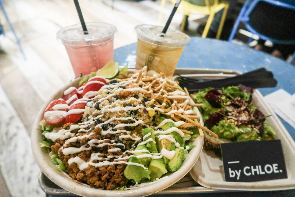 Caption: A photo of a vegetarian quinoa bowl topped with avocado, fake meat, tortilla strips, and tomatoes drizzled with a white crema sauce alongside a piece of avocado toast and two soft drinks in plastic cups from by CHLOE in New York City. (Local Guide Marco Togni)