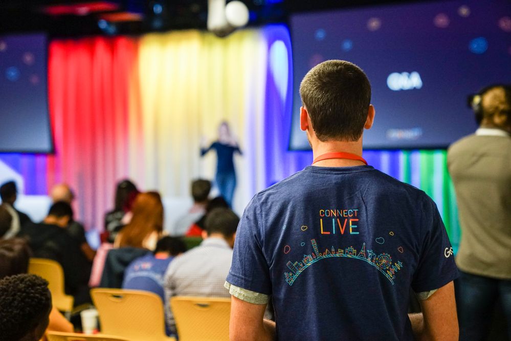 Caption: A photo of the back of a Googler wearing a Connect Live 2018 T-shirt while watching Jen Fitzpatrick, VP of Google Maps, deliver a keynote.
