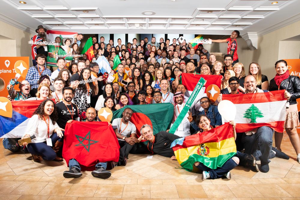 Caption: A group photo of Local Guide attendees at Connect Live 2018 holding up flags from their home countries as well as Local Guides props.