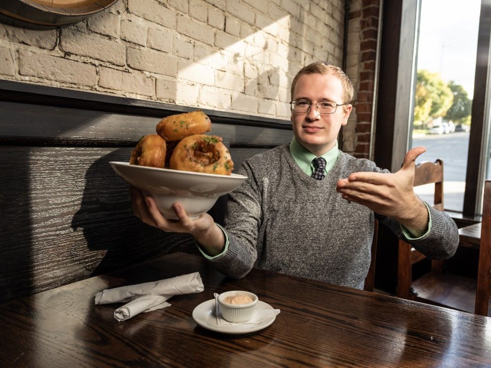 Caption: A photo of Local Guide Tyler Groenendal holding up a bowl of onion rings in one hand and pointing to it with other at a table at Graydon’s Crossing in Grand Rapids, Michigan. (Brian Kelly)