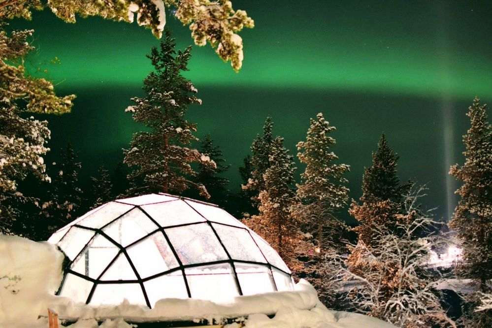 Caption: A photo of a glass igloo at Kakslauttanen Arctic Resort in Sodankylä, Finland surrounded by snow, trees, and the Northern Lights. (Local Guide Gareth Powell)
